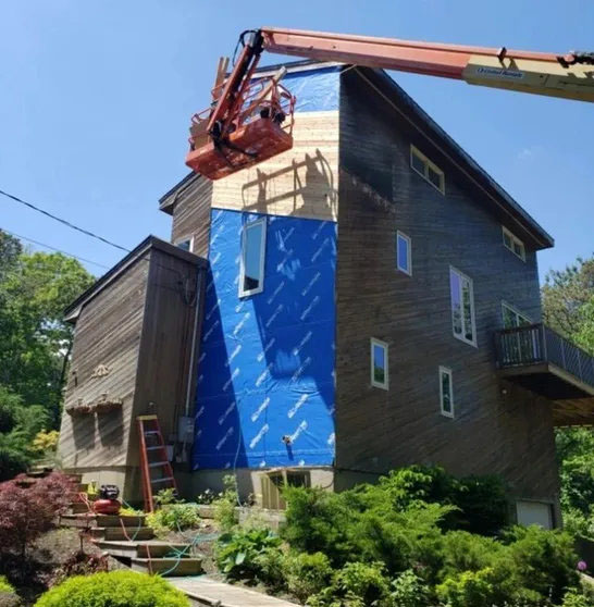 A construction worker uses a cherry picker to install siding on the upper section of a house, partially covered in blue tarps.