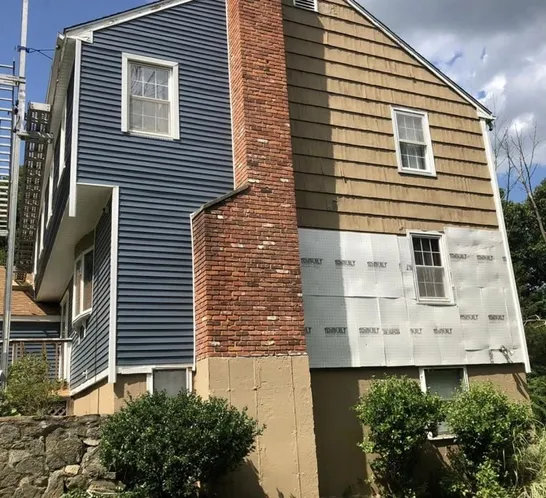 A two-story house featuring a mix of blue siding, brown shingles, exposed brick, and unpainted wall preparations, with greenery below.