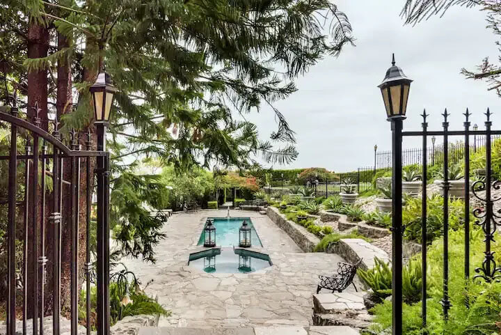 About Us - A serene garden pathway leads to a tranquil pool, surrounded by lush greenery and elegant lanterns under a cloudy sky.