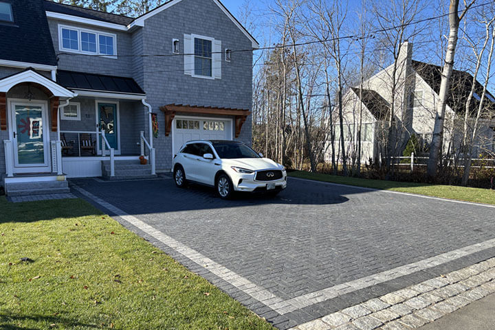About Us - White SUV parked on a cobblestone driveway in front of a gray house surrounded by trees, with blue sky above.