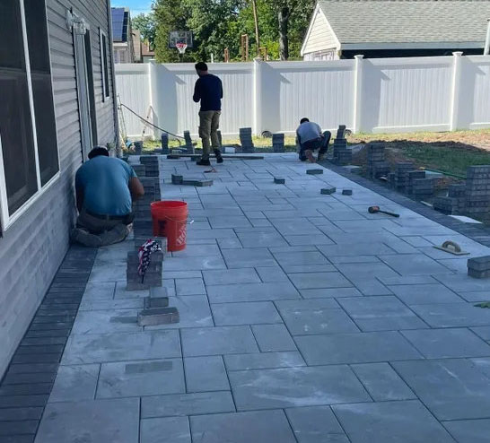 Workers installing pavers on a patio, surrounded by scattered bricks and tools, with a white fence and trees in the background.