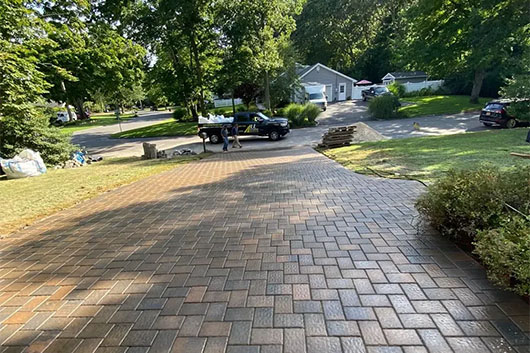 A newly paved brick driveway curves through a landscaped yard, leading to parked cars and homes in a peaceful neighborhood.