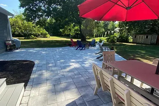 A spacious outdoor patio with a large table under a red umbrella, surrounded by colorful chairs and a lush green lawn.