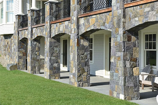 Stone pillars with arches support a balcony overlooking a manicured lawn, featuring white doors and wooden chairs beneath a blue sky.