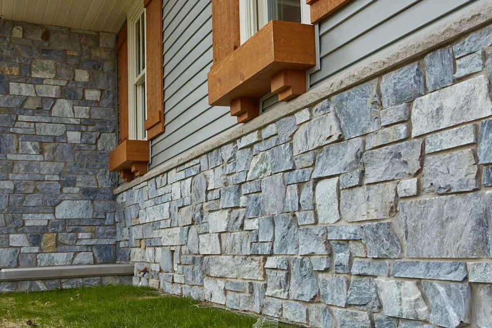 Close-up of a gray stone facade beneath wooden window trim, showcasing textured stone work and green grass.