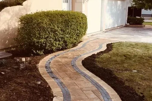 A winding brick walkway with blue accents curves through a landscaped yard, leading to a garage beside a lush green hedge.