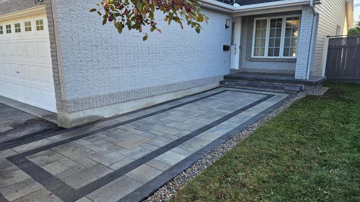 Driveway with patterned pavers in gray and black, leading to a house with a porch and large windows. Green grass on the side.