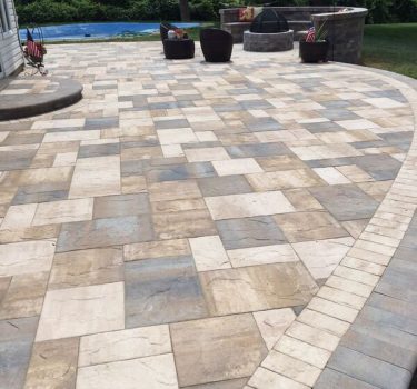 A landscaped patio featuring interlocking stone pavers in beige and gray, with seating and a fire pit, surrounded by greenery.