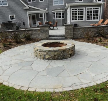 A stone fire pit surrounded by a circular flagstone patio, with a gray house and outdoor seating in the background.