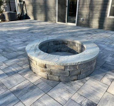A circular stone fire pit surrounded by gray paved patio tiles, set against a backdrop of a house and fenced yard.