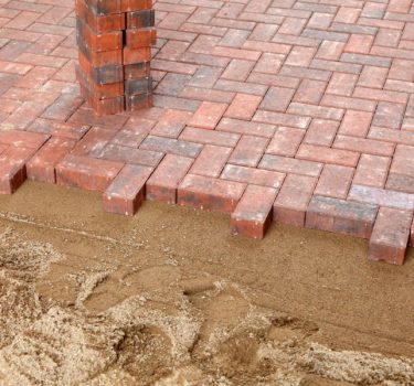 Close-up of a brick patio with herringbone pattern, featuring a sandy edge where the bricks meet the ground.