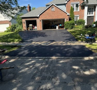 A freshly paved driveway in front of a red brick house with a garage; tools and a wheelbarrow sit nearby on concrete.