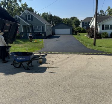 Two workers outdoors near a freshly paved driveway; one fills a wheelbarrow while a truck is unloading materials in the background.