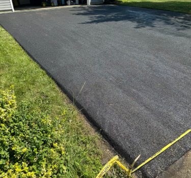 Freshly paved black asphalt driveway beside a well-kept lawn and a garage, marked with yellow caution tape.