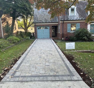 A renovated home featuring a new stone driveway, blue garage door, and landscaped yard with autumn leaves. A sign for a local contractor is visible.