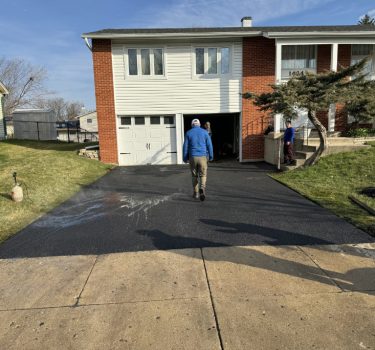 A person walks on a newly paved driveway in front of a suburban home, with another person working nearby and blue sky above.