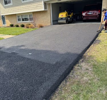 A worker spreads fresh asphalt on a driveway in front of a two-story house, with a parked red car and paving equipment nearby.