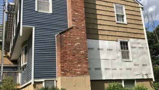 A two-story house featuring a mix of blue siding, brown shingles, exposed brick, and unpainted wall preparations, with greenery below. A two-story house featuring a mix of blue siding, brown shingles, exposed brick, and unpainted wall preparations, with greenery below.