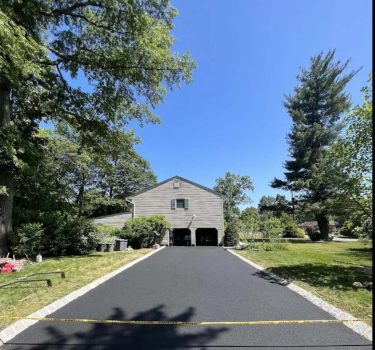 A newly paved black asphalt driveway leads to a two-garage house surrounded by lush greenery under a clear blue sky.