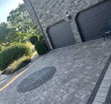 A modern home with a stone exterior, featuring two dark garage doors, a circular paved design on the driveway, and green hedges.