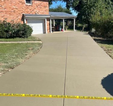 Freshly poured concrete driveway with caution tape, leading to a brick house and a covered porch, surrounded by grassy areas.