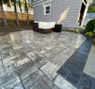 Paved patio featuring large, gray stone tiles with a wicker sofa set, surrounded by trees and a house in the background.