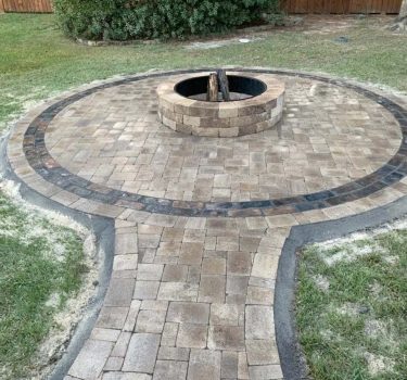 A circular stone patio surrounds a fire pit, with a winding pathway leading through a grassy backyard and wooden fence in the background.
