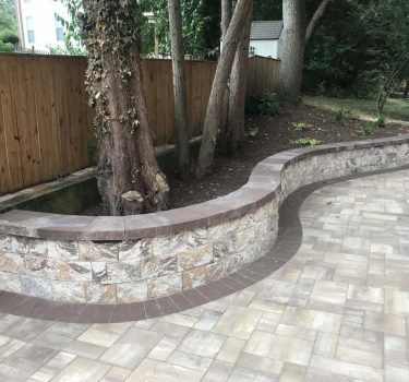 A curved stone bench surrounds a tree in a landscaped yard, bordered by a wooden fence and paved with light-colored bricks.