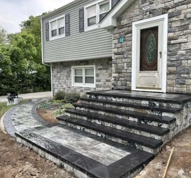 A new stone entrance with wide black steps and a curved walkway leads to a house with gray siding and a decorative front door.