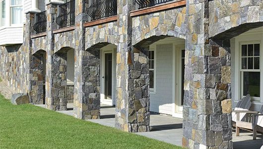 Stone pillars with arches support a balcony overlooking a manicured lawn, featuring white doors and wooden chairs beneath a blue sky. Stone pillars with arches support a balcony overlooking a manicured lawn, featuring white doors and wooden chairs beneath a blue sky.