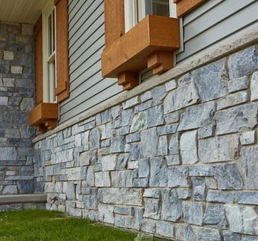 Close-up of a gray stone facade beneath wooden window trim, showcasing textured stone work and green grass.