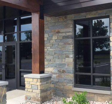 A modern stone-clad entrance features wooden beams, large windows, and a stylish door, set against a landscaped area.