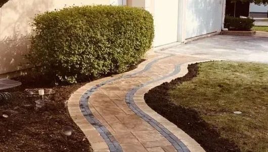 A winding brick walkway with blue accents curves through a landscaped yard, leading to a garage beside a lush green hedge. A winding brick walkway with blue accents curves through a landscaped yard, leading to a garage beside a lush green hedge.
