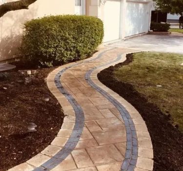 A winding brick walkway with blue accents curves through a landscaped yard, leading to a garage beside a lush green hedge.