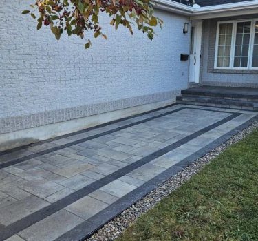 Driveway with patterned pavers in gray and black, leading to a house with a porch and large windows. Green grass on the side.