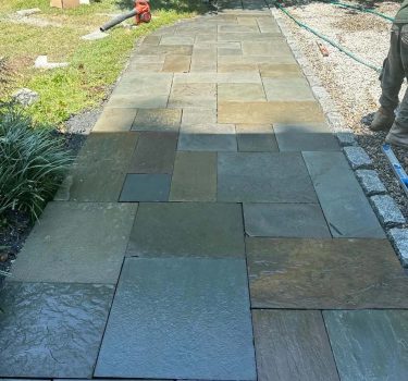 A newly laid stone pathway with various shades of slate tiles, surrounded by grass and garden tools in a well-kept outdoor area.