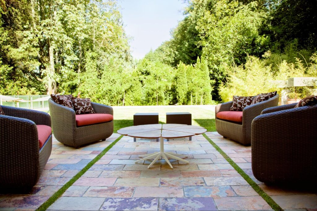 A cozy outdoor patio with wicker chairs, red cushions, and a circular coffee table surrounded by lush greenery and stone flooring.