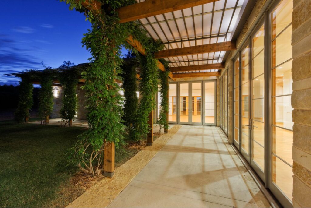 A modern outdoor walkway with a pergola featuring stone-textured flooring, lush green climbing plants, and glass-paneled doors leading into a well-lit interior.