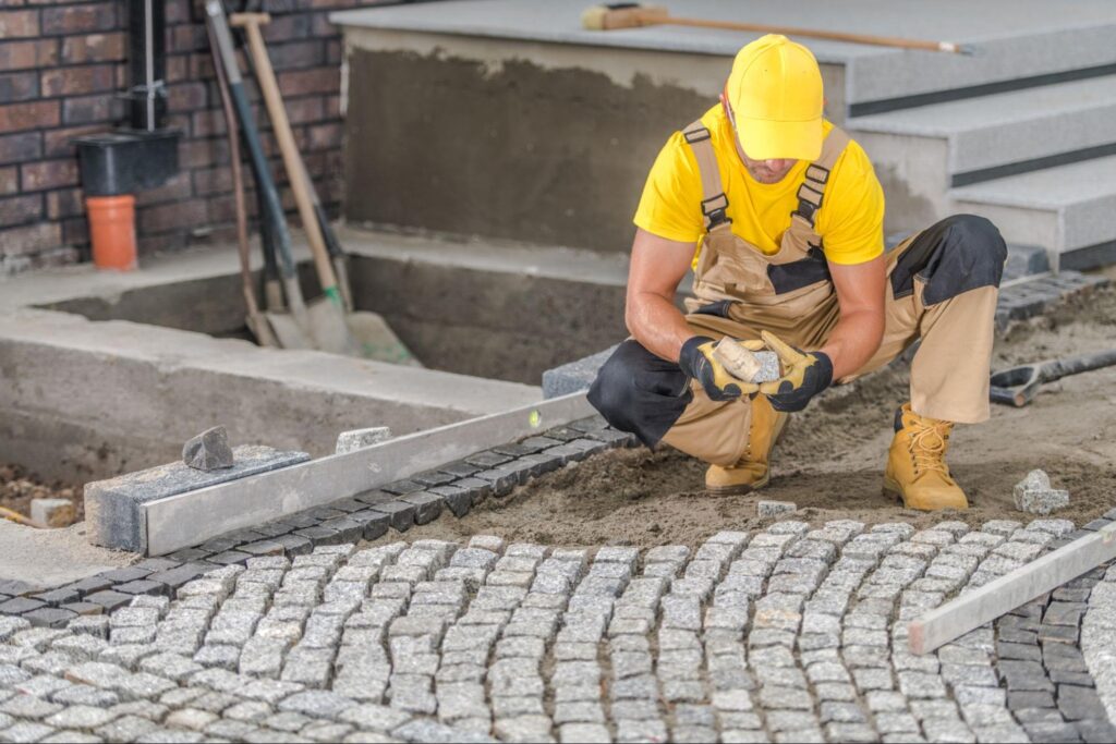A contractor in a yellow uniform lays paving stones on the driveway.